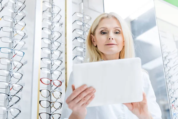 Professional female ophthalmologist working with digital tablet in optics with glasses on shelves — Stock Photo