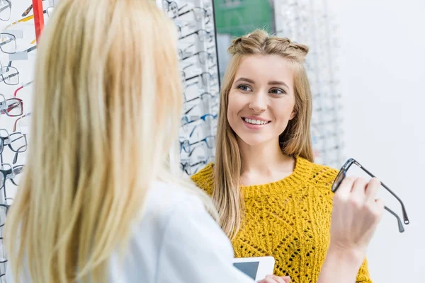 Oculist helping beautiful client to choose eyeglasses in optics — Stock Photo