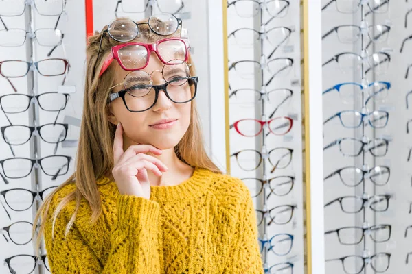 Joven chica pensativa la elección de las gafas en óptica - foto de stock