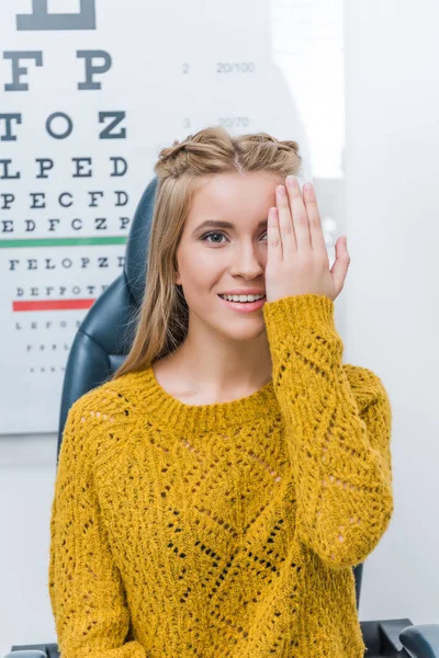 Young female patient with eye test in clinic with eye chart behind — Stock Photo