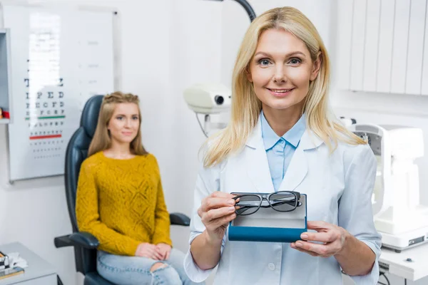 Optician in white coat holding eyeglasses while patient sitting behind — Stock Photo