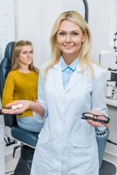 Optometrista en bata blanca sujetando anteojos y lente de contacto, paciente sentado detrás - foto de stock