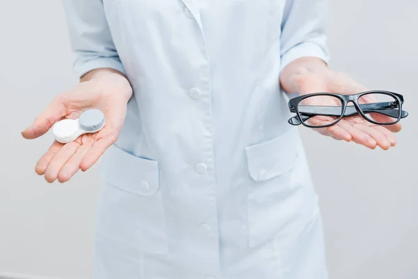 Vista recortada del optometrista en capa blanca que sostiene las gafas y la lente de contacto, aislado en blanco - foto de stock