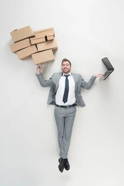 Vista aérea del empresario sosteniendo cajas de cartón y utilizando un ordenador portátil aislado en gris - foto de stock