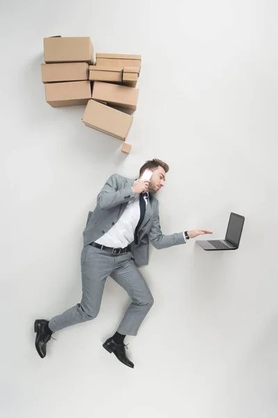 Overhead view of businessman with cardboard boxes above head talking on smartphone and using laptop isolated on grey — Stock Photo