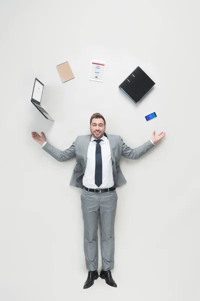 Overhead view of smiling businessman with outstretched arms and office supplies above head looking at camera isolated on grey — Stock Photo