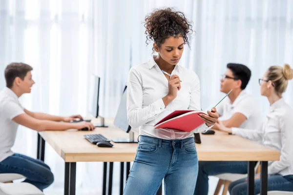 Giovane ragazza studentessa africana americana pensierosa che scrive sul quaderno in classe con gli amici — Foto stock