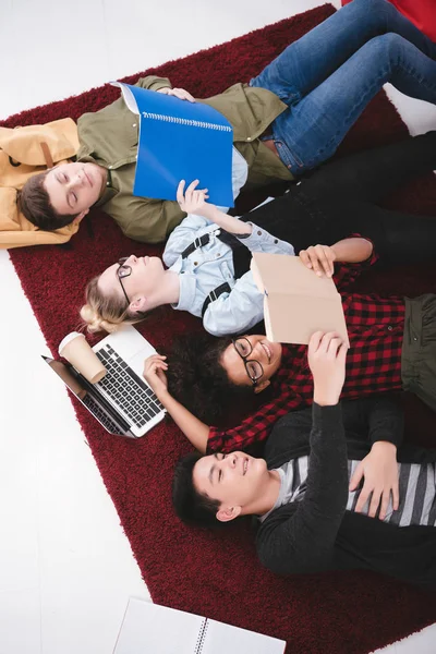 Jeunes étudiants adolescents couchés sur le tapis avec des cahiers et un ordinateur portable — Photo de stock