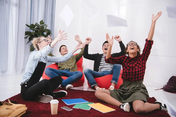 Jóvenes estudiantes felices sentados y lanzando papeles al aire - foto de stock