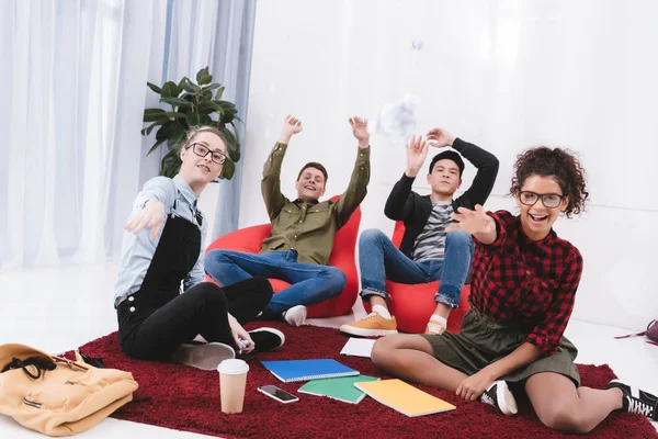 Jóvenes estudiantes felices sentados y arrojando papel en la cámara - foto de stock