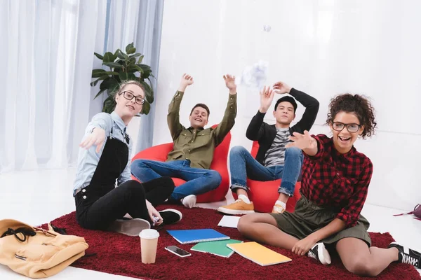 Jovens estudantes felizes sentados e jogando papel na câmera — Fotografia de Stock