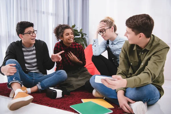 Young laughting teenagers sitting on carpet, studying and pointing on girl — Stock Photo