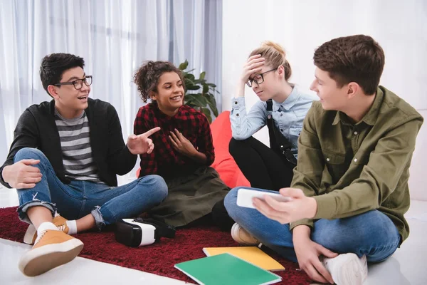 Junge lachende Teenager, die auf Teppich sitzen, studieren und auf Mädchen zeigen — Stockfoto