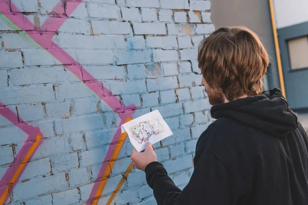 Street artist looking at sketch and painting colorful graffiti on wall — Stock Photo