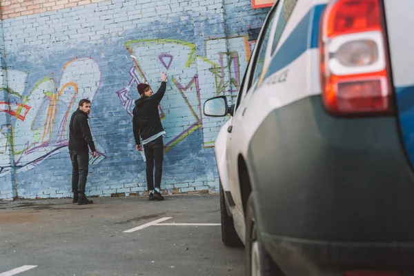 Back view of vandals painting graffiti on wall, police car on foreground — Stock Photo