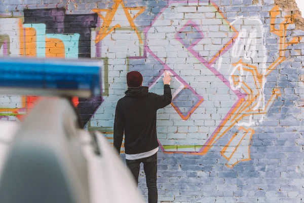 Back view of vandal painting graffiti on wall, police car on foreground — Stock Photo