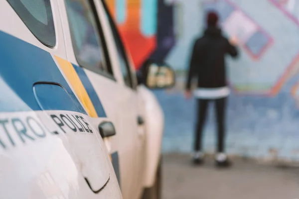 Back view of vandal painting graffiti on wall, police car on foreground — Stock Photo