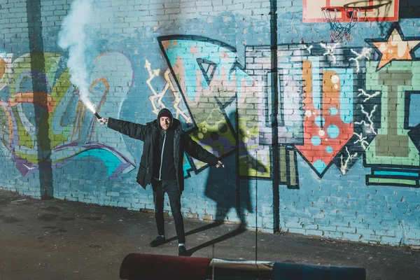 Mann hält Rauchbombe in der Hand und steht nachts mit Graffiti gegen Wand — Stockfoto