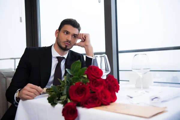 Giovane in giacca e cravatta con bouquet di rose rosse in attesa di fidanzata nel ristorante — Foto stock