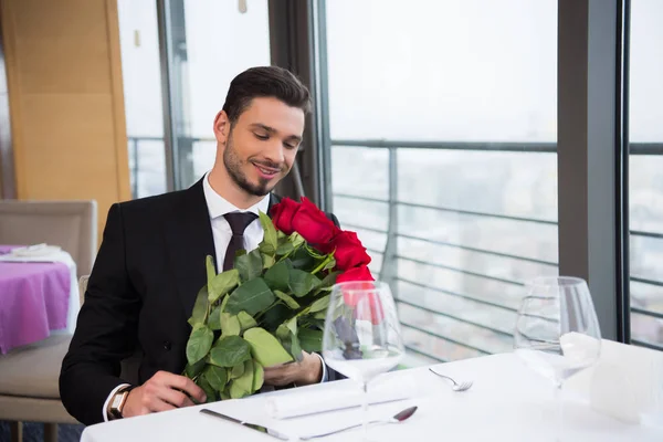 Улыбающийся мужчина в костюме с букетом красных роз ждет свою девушку в ресторане — стоковое фото