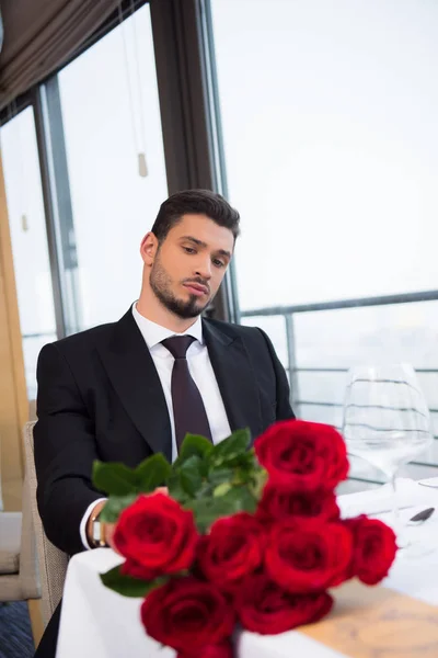 Portrait of young man in suit with bouquet of red roses waiting for girlfriend in restaurant — Stock Photo
