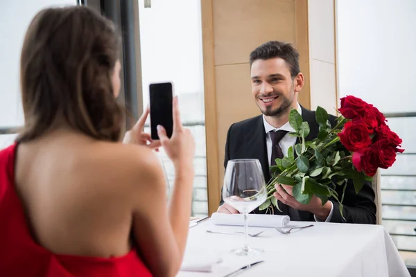 Vue partielle de petite amie prendre des photos de petit ami souriant avec bouquet de roses au restaurant pendant la date romantique — Photo de stock