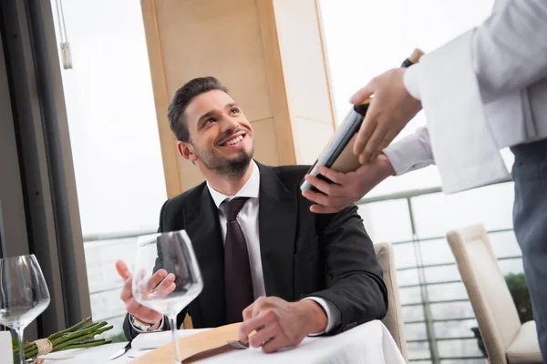 Частичный вид улыбающегося человека, смотрящего на официанта с бутылкой вина в ресторане — стоковое фото