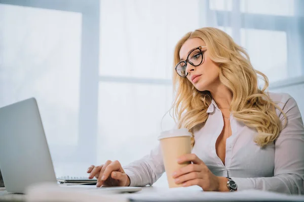 Beautiful businesswoman working at laptop in office and holding coffee in paper cup — Stock Photo