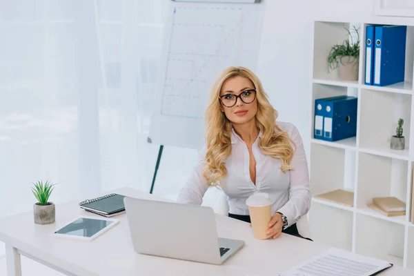 Hermosa mujer de negocios sentada en la mesa con el ordenador portátil y la celebración de taza de café desechable - foto de stock