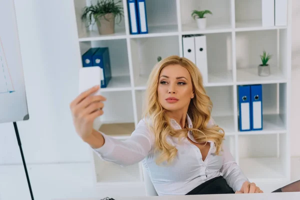 Serious sexy businesswoman taking selfie at working place with smartphone — Stock Photo