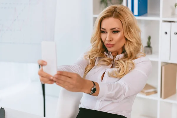 Sexy mujer de negocios tomando selfie en el lugar de trabajo con teléfono inteligente - foto de stock