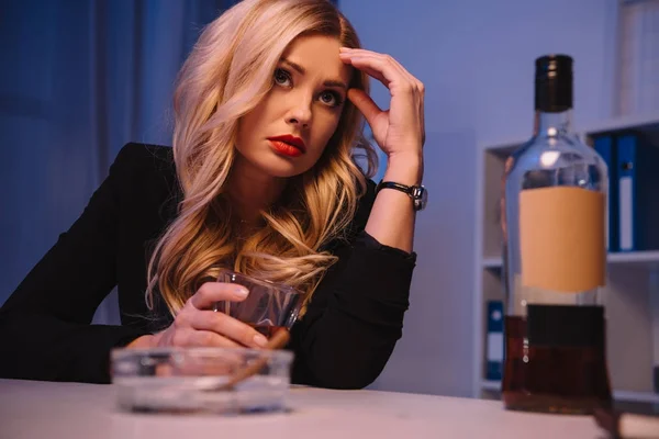 Mujer de negocios rubia cansada sentada en la mesa con un vaso de whisky - foto de stock