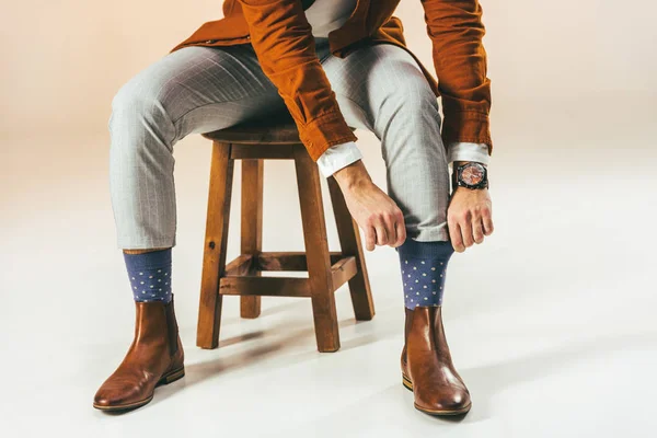 Обрізаний знімок стильного чоловіка, що зв'язує шкарпетки, сидячи на дерев'яному стільці, на бежевому — стокове фото