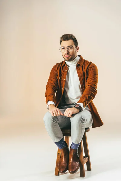 Nachdenklicher junger Mann in modischer Kleidung auf einem Holzstuhl sitzend, auf beige — Stockfoto