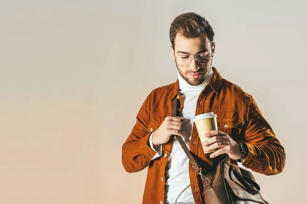 Ritratto di uomo alla moda con borsa guardando monouso tazza di caffè in mano isolato su beige — Foto stock