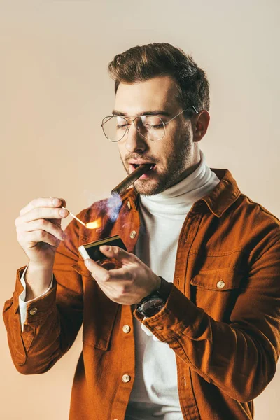 Porträt eines stilvollen Mannes mit Brille, der Zigarre isoliert auf Beige anzündet — Stockfoto