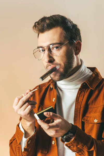 Портрет стильного чоловіка в окулярах, що освітлює сигару ізольовано на бежевому — стокове фото