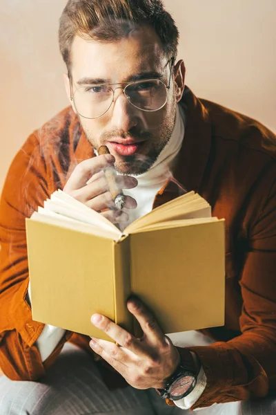 Porträt eines jungen Mannes mit Zigarre und Buch in der Hand, der isoliert in die beige Kamera blickt — Stockfoto