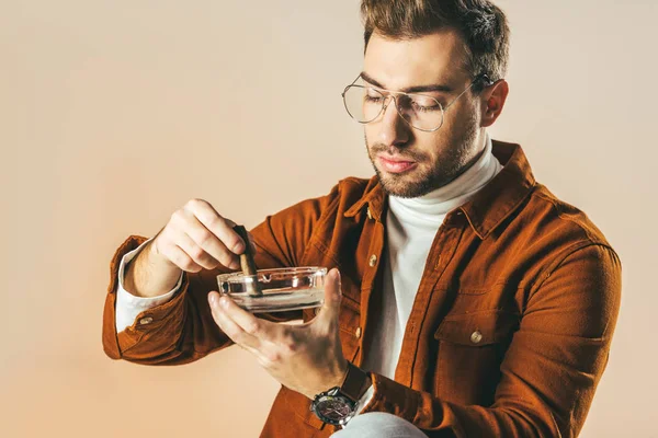 Portrait of man in eyeglasses putting out a cigar in ashtray isolated on beige — Stock Photo