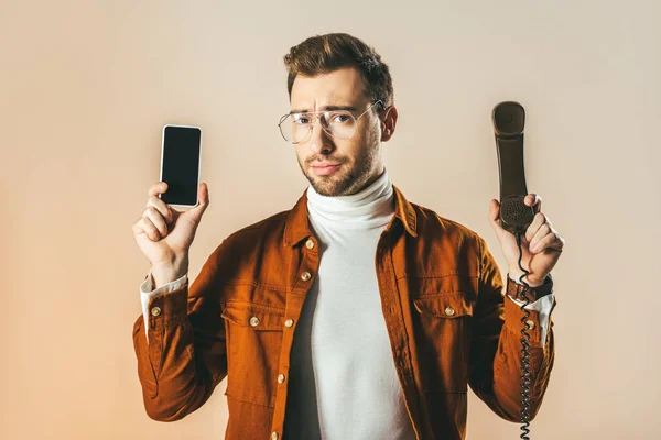 Portrait of handsome man showing smartphone and telephone tube in hands isolated on beige — Stock Photo