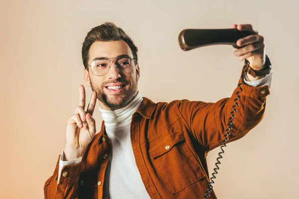 Portrait of smiling man showing peace sign while pretending taking selfie in telephone tube isolated on beige — Stock Photo