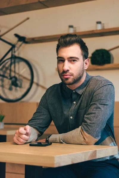 Ritratto dell'uomo pensieroso che guarda la macchina fotografica mentre siede a tavola con lo smartphone nel caffè — Foto stock