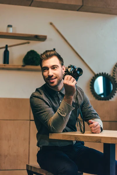 Retrato de hombre sonriente con cámara fotográfica en la mano sentado en la mesa en la cafetería - foto de stock
