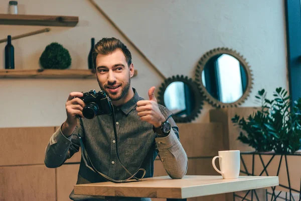 Porträt eines jungen Mannes mit Fotokamera, die Daumen hoch im Café zeigt — Stockfoto