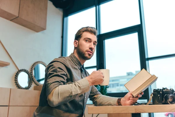 Vista laterale dell'uomo con tazza di caffè e libro in mano seduto a tavola nel caffè — Foto stock