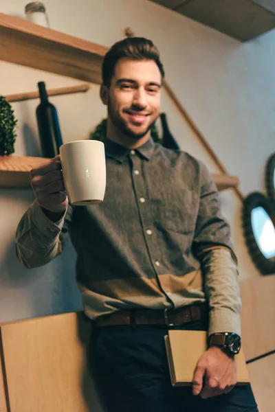 Enfoque selectivo del hombre sonriente con taza de café y libro en las manos en la cafetería - foto de stock