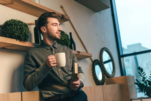 Портрет улыбающегося мужчины с чашкой кофе и книгой в руках в кафе — стоковое фото