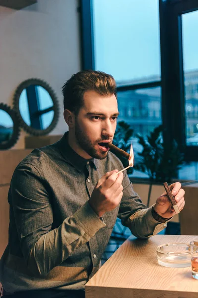 Portrait of young man lighting up cigar while sitting at table in cafe — Stock Photo