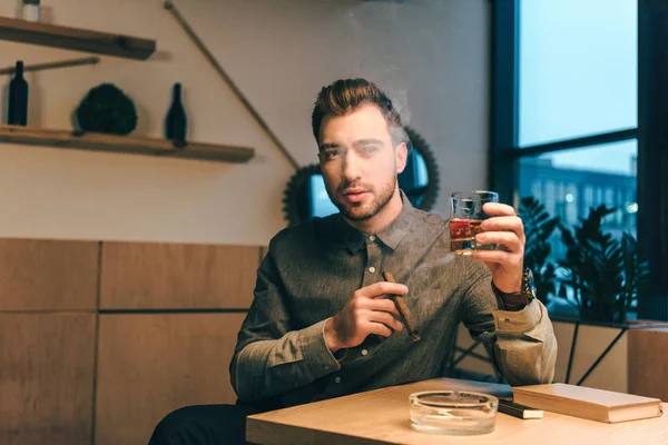 Retrato de hombre joven con vaso de coñac y cigarro en las manos sentado a la mesa en la cafetería - foto de stock