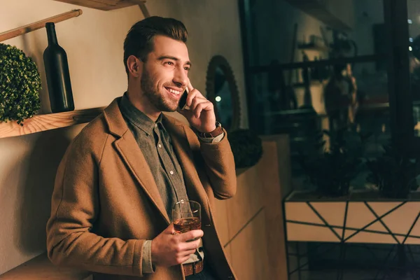 Side view of smiling man with glass of whiskey talking on smartphone in cafe — Stock Photo